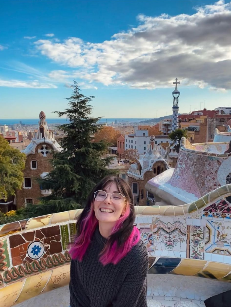 alt="Retrato de una mujer joven sonriente con el pelo oscuro y puntas rosas, sentada en el Park Güell de Barcelona con la ciudad de fondo bajo un cielo azul."