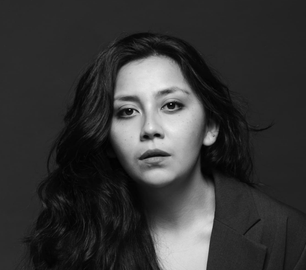 alt="Fotografía artística en blanco y negro de una autora con cabello largo y ondulado, mirando seriamente a la cámara con una chaqueta oscura."