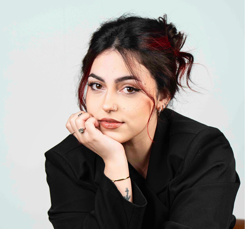 alt="Retrato de una joven autora con cabello oscuro y mechones rojos, posando con la barbilla apoyada en su mano y vistiendo una chaqueta negra."